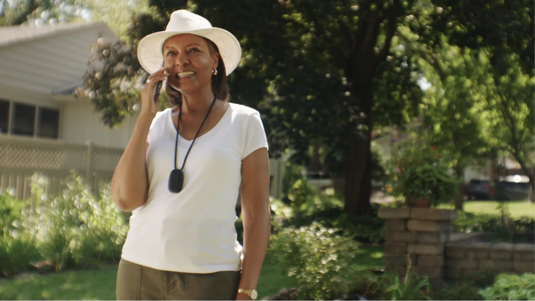 A woman dressed in a white shirt and hat, talks on the phone and smiles in her garden while wearing a Personal Emergency Response pendant.