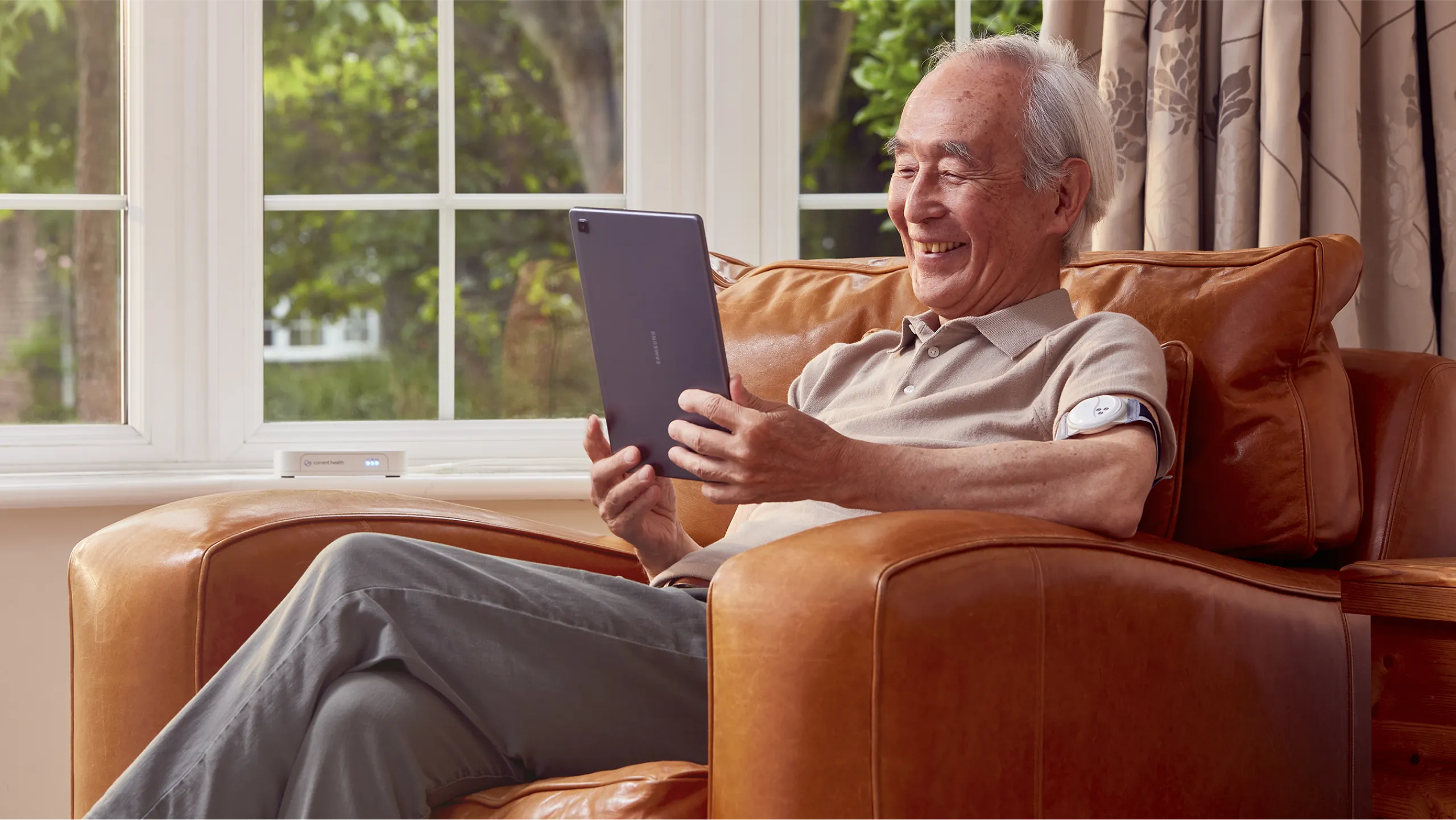 An smiling elderly man wears a Current Health Continuous Vital Sign Monitor on his arm, while  looking at his tablet.