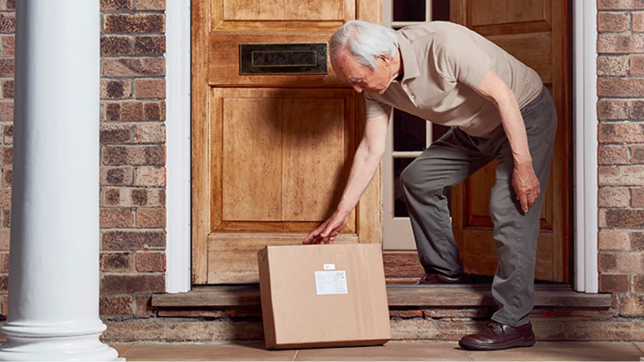 An elderly man picks up a package at his front door.