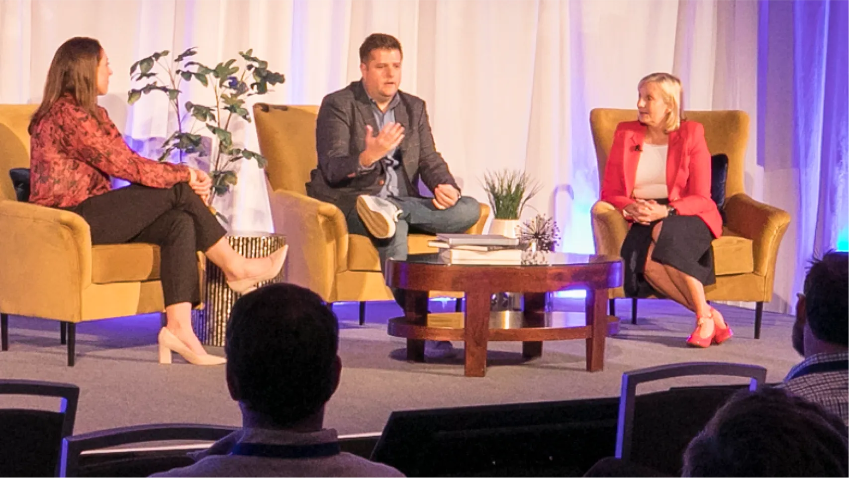 Three business professionals sit on stage and chat in front of an audience.