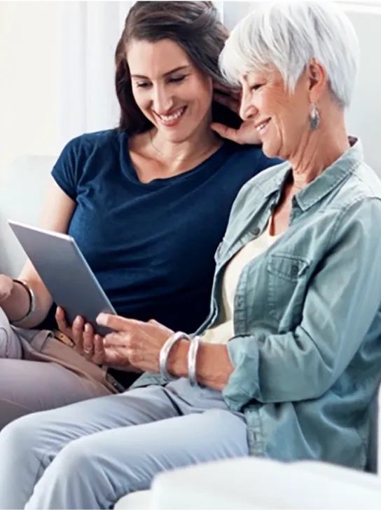 A mother and adult daughter smiling as they look at a tablet.