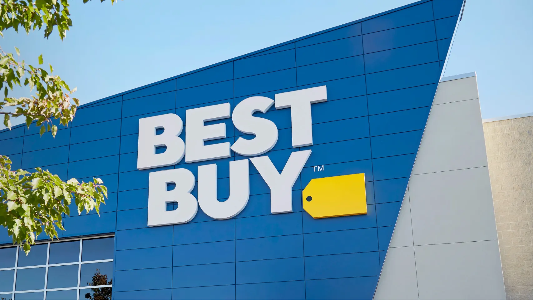 The Best Buy logo on the front of a Best Buy store.  “Best Buy” is in large white text with a yellow tag next to it.