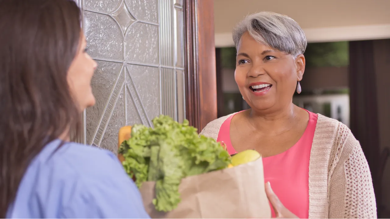 A woman delivers vegetables to another woman at her front door.