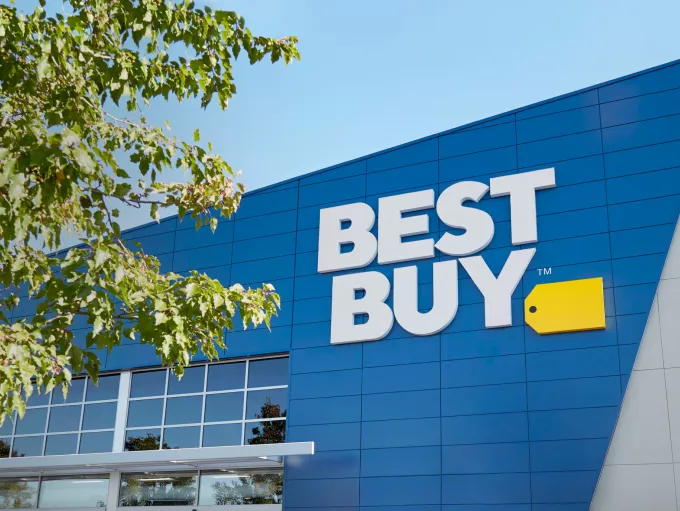 The Best Buy logo on the front of a Best Buy store.  “Best Buy” is in large white text with a yellow tag next to it.