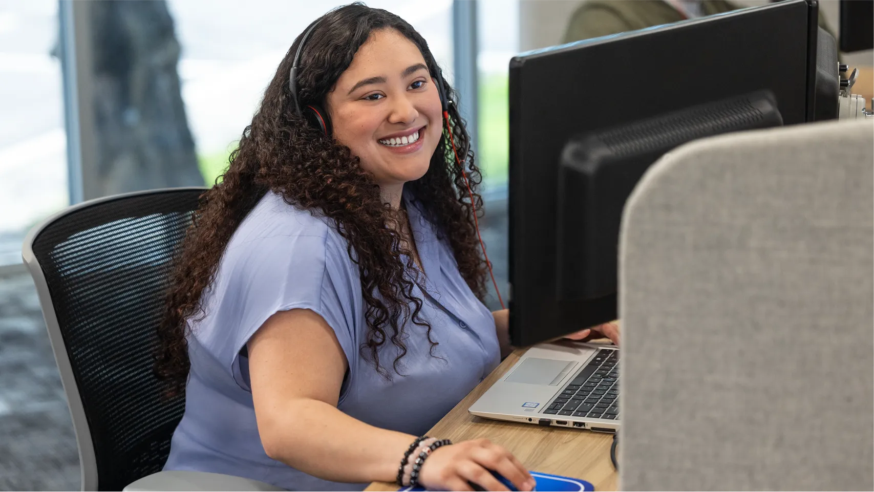 A customer care agent with long curly hair sits at a desk with a headset and smiles into the computer.